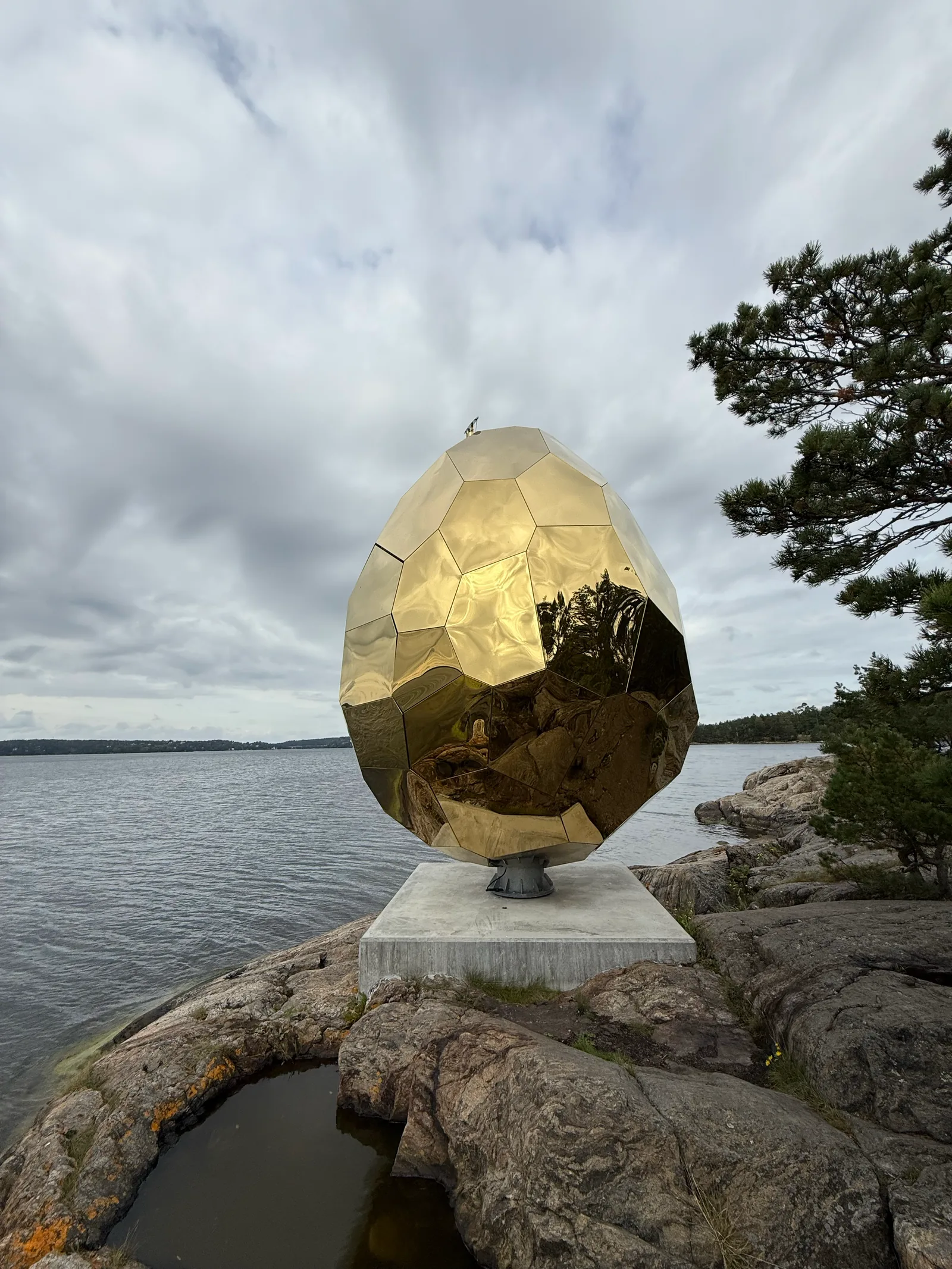 Goldenes, facettiertes Ei aus Spiegelblech auf dem Fels am Wasser bei Artipelag