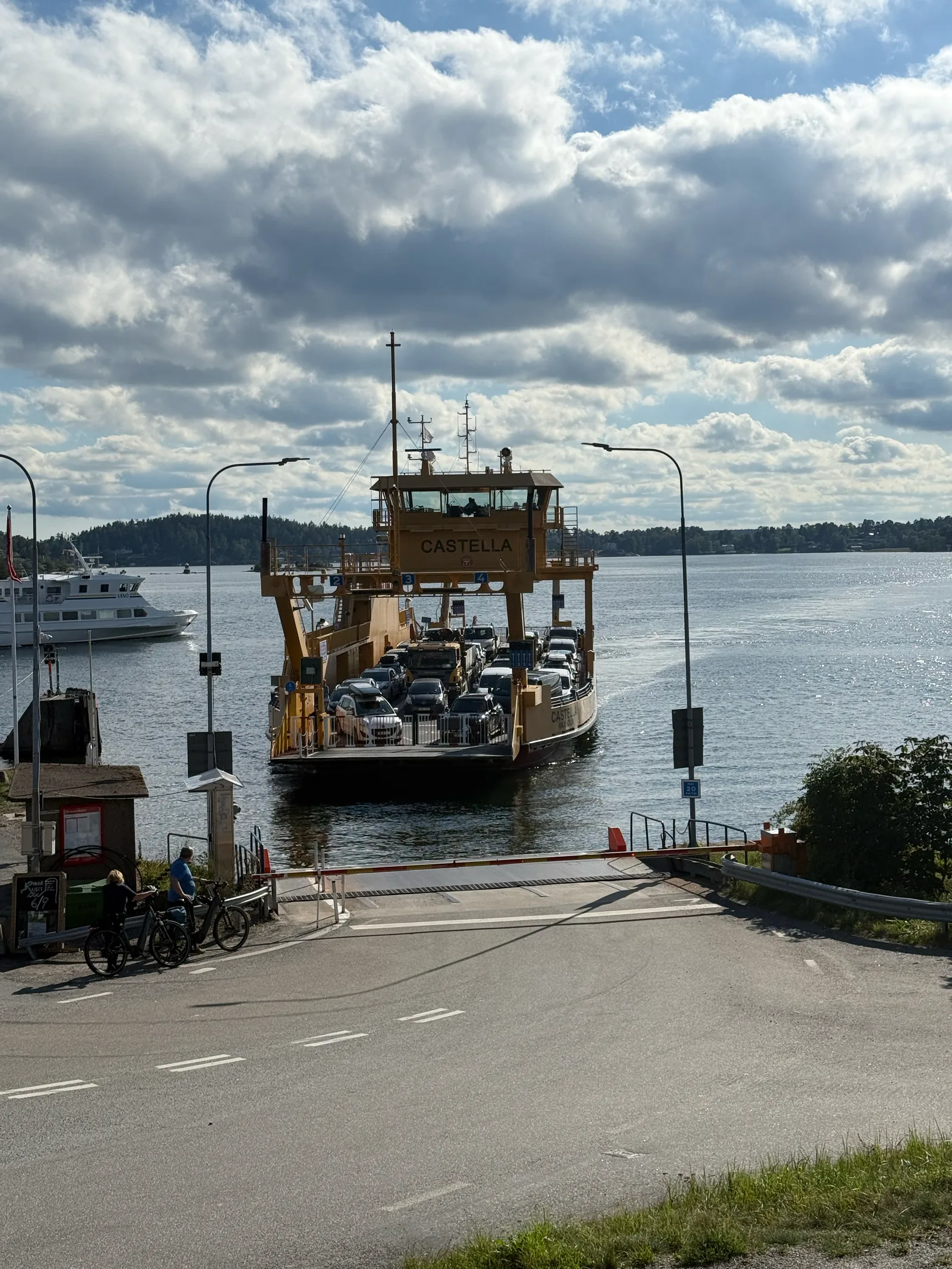 Gelbe Autofähre legt am Anleger bei Vaxholm an, Radfahrer warten an der Schranke