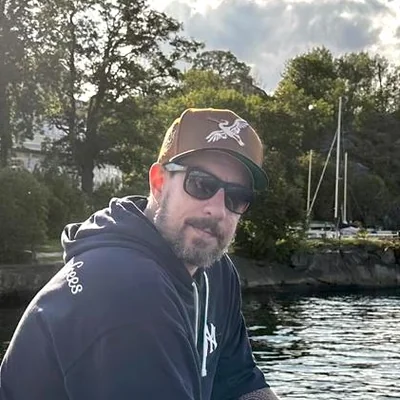 Martin Höllinger mit Kappe und Sonnenbrille auf einem Boot, dahinter Ufer und Masten