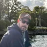 Martin Höllinger mit Kappe und Sonnenbrille auf einem Boot, dahinter Ufer und Masten