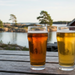 Zwei gefüllte Craft-Beer-Gläser auf einem Holztisch mit unscharfem Schärenhafen im Hintergrund