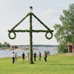 Mit Birkengrün und Wildblumen geschmückter Midsommarstång mit zwei Ringen auf einer Wiese am Wasser, Menschen im Tanzkreis in der Ferne